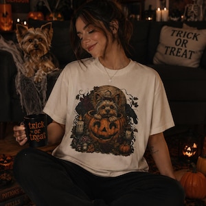 Puede incluir: Una camiseta de color crema con un estampado gráfico de un Yorkshire Terrier sentado dentro de una calabaza tallada, rodeado de hojas de otoño, setas y velas. La camiseta se combina con pantalones negros y una taza negra con el texto Trick or Treat.