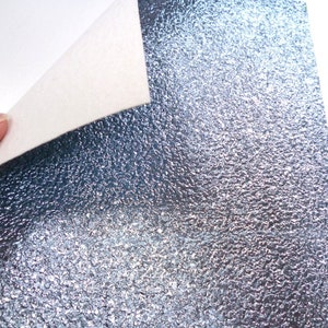 May include: A sheet of silver glitter fabric with a textured, bumpy surface. The fabric has a subtle blue hue.