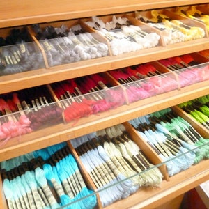 Puede incluir: Una estantería de madera con varios cajones llenos de carretes de hilo de bordar de colores. El hilo está organizado por colores y dispuesto en filas.