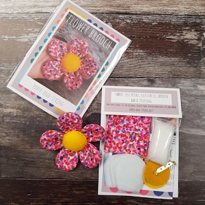 May include: A handmade flower brooch kit with a finished flower. The flower has pink petals with colorful speckles and a yellow center. The kit includes fabric, felt circles, a brooch bar, and stuffing. The packaging reads "FLOWER BROOCH" and "PAPER-AND-STRING".