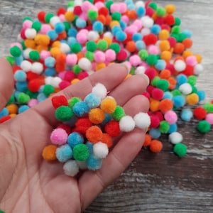 Puede incluir: Una mano sosteniendo una variedad de pompones coloridos en una variedad de colores, incluyendo rojo, naranja, amarillo, verde, azul y blanco.