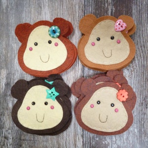 May include: Four felt monkey face coasters in shades of brown and tan. Each coaster features a smiling face with pink cheeks and a decorative button. The buttons are in the shape of a flower, star, and heart.