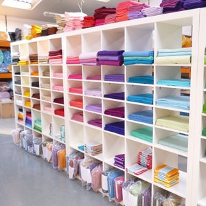 May include: A white shelf unit filled with stacks of colorful felt fabric in various shades of pink, purple, blue, green, yellow, and orange. The felt is organized by color and neatly stacked on the shelves.
