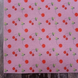 May include: Pink fabric featuring a repeating pattern of red cherries and green leaves. The cherries are arranged in clusters of varying sizes, creating a lively design. The fabric has a soft, matte finish.