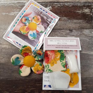 May include: A handmade flower brooch kit with fabric, felt petals, felt circles, brooch bar, and stuffing. The kit includes a finished flower brooch with a yellow center and colorful petals. The packaging reads "FLOWER BROOCH" and "PAPER-AND-STRING."