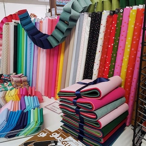 May include: A shop display featuring a wide array of fabrics and craft materials. Rolls of patterned and plain fabric are arranged vertically. Stacks of felt in different colours are present, along with a felt garland and other craft supplies.