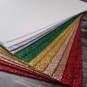 May include: A stack of glitter paper in various colours, including white, silver, green, gold, pink, and red.