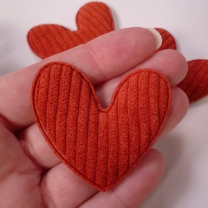 May include: Three red felt hearts with a ribbed texture. The hearts are arranged in a row, with the largest heart in the centre.