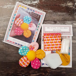 May include: A craft kit for making a flower brooch. The kit includes fabric, felt petals, felt circles, a brooch bar, and stuffing. The flower brooch is made of colorful fabric with polka dots and other patterns. The packaging reads "FLOWER BROOCH" and "PAPER-AND-STRING".