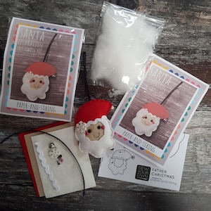 Könnte beinhalten: Ein weihnachtliches Filz-Nähset mit einem Weihnachtsmann-Design. Das Set enthält Filzteile, eine Nadel, Faden und Füllwatte. Die Verpackung trägt den Text "SANTA DECORATION FELT SEWING KIT" und "PAPER-AND-STRING".