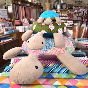 May include: Two plush turtle toys stacked vertically. The larger bottom turtle features a multi-coloured floral patchwork shell and beige fleece limbs. The smaller top turtle has a bright blue head and a green shell patterned with pink flowers.