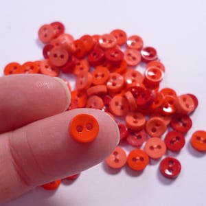 Peut inclure: Un assortiment de petits boutons ronds dans des tons de rouge et d'orange. Un bouton orange vif à deux trous est tenu entre les doigts, avec de nombreux autres boutons dispersés en arrière-plan. Ces boutons sont parfaits pour la couture et les travaux manuels.