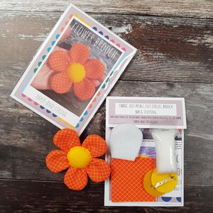 May include: A flower brooch craft kit with orange and yellow fabric. The kit includes fabric, felt petals, felt circles, a brooch bar, and stuffing. The packaging features the text "FLOWER BROOCH" and "PAPER-AND-STRING".