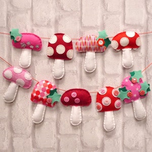May include: A garland of felt mushroom decorations in shades of pink and red with white polka dots. The mushrooms are strung together with green felt leaves and red buttons.