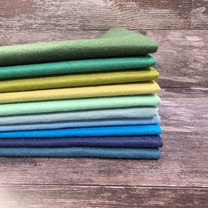 May include: A pile of felt fabric in a spectrum of greens and blues. The colours transition from light green to dark navy, forming a gradient. The fabric has a soft texture and a subtle sheen, suggesting a high-quality material.