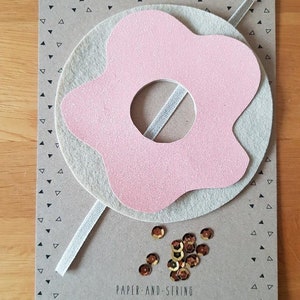 May include: A pink felt donut with a hole in the centre, surrounded by a beige felt circle. The donut is decorated with gold glitter. The image also includes a white ribbon and gold sequins.