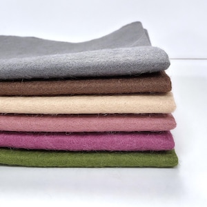 Op de afbeelding: Een stapel van zes rechthoekige viltvellen in verschillende kleuren, waaronder grijs, bruin, beige, lichtroze, donkerroze en olijfgroen. Elk vel heeft een zachte, vezelige textuur en is netjes op elkaar gestapeld.