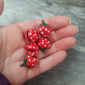 Könnte beinhalten: Fünf rote, erdbeerförmige Glocken mit weißen Punkten und grünen Stielen. Die Glocken werden in einer Hand gehalten, wodurch ihre leuchtenden Farben und ihr verspieltes Design hervorgehoben werden. Diese dekorativen Glocken könnten für Bastelarbeiten oder Feiertagsdekorationen verwendet werden.