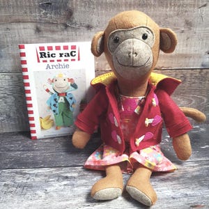 May include: A plush monkey doll wearing a red jacket with pink accents and a floral dress. The doll is next to a book titled "Ric rac Archie" with a cartoon monkey on the cover. The background is a wooden surface.