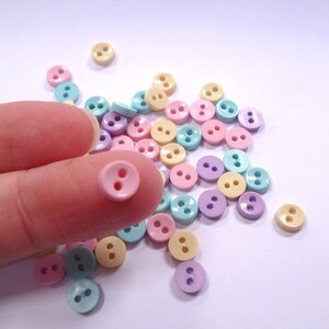 Può includere: Una collezione di bottoni color pastello in rosa, giallo, blu e viola. I bottoni sono piccoli e hanno due fori.
