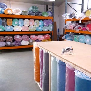 May include: A workshop with shelves full of colorful fabric rolls in various sizes. The fabric rolls are wrapped in clear plastic and are organized by color. A wooden table with a tape dispenser and a silver iron sits in the center of the room.