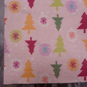 May include: Pink felt fabric with a repeating pattern of colorful Christmas trees and snowflakes. The trees are in shades of green, red, orange, and yellow. The snowflakes are in shades of pink, orange, and blue.