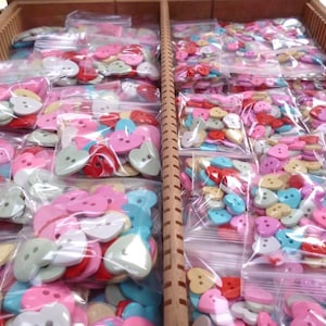 Puede incluir: Una gran colección de botones con forma de corazón en varios colores, incluyendo rosa, rojo, azul, verde y blanco. Los botones están empaquetados en bolsas de plástico transparentes.