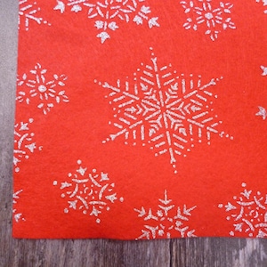 May include: Red felt fabric with a white snowflake pattern. The snowflakes are made of glitter and have a delicate design.