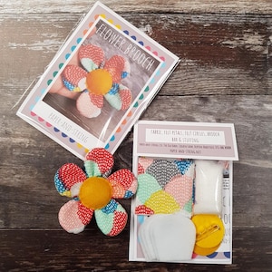 May include: A flower brooch craft kit with a finished brooch. The brooch is a colorful fabric flower with a yellow center. The kit includes fabric, felt petals, felt circles, a brooch bar, and stuffing. The packaging reads "FLOWER BROOCH" and "PAPER-AND-STRING."