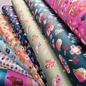 May include: A stack of folded fabric bolts featuring various patterns including owls, hedgehogs, bears, mushrooms, and floral motifs in colors of pink, dark green, olive green, navy blue, yellow, and light blue with a woven texture.