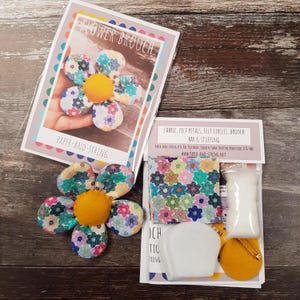 May include: A flower brooch kit with a finished brooch. The kit includes fabric, felt petals, felt circles, brooch bar, and stuffing. The finished brooch is a flower with a yellow center and patterned petals. The packaging reads "FLOWER BROOCH" and "PAPER-AND-STRING."