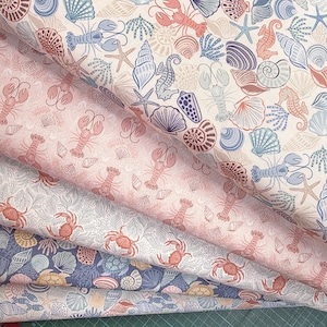 May include: Close-up of several fabric samples with a nautical theme. The fabrics feature repeating patterns of seashells, starfish, lobsters, and seahorses in shades of blue, coral, and pink on a white background. The fabrics are layered.