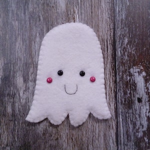 May include: A white felt ghost with a smiling face, black eyes, and pink cheeks. The ghost is on a wood background.