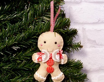 May include: A gingerbread man Christmas ornament made of felt. The ornament is beige with a red and white bow and red buttons. The ornament has a white trim and a red and white striped ribbon for hanging.