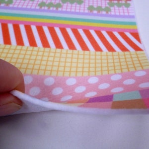 May include: A close-up of a colorful patterned fabric with a white backing. The fabric features a variety of geometric shapes and patterns, including stripes, polka dots, and checkered designs. The fabric is folded over to reveal the white backing.