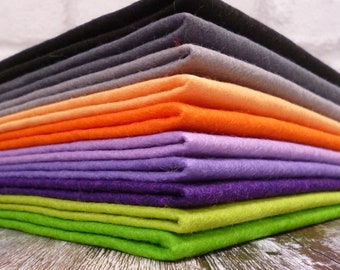 Op de afbeelding: Een stapel viltstof in verschillende kleuren, waaronder zwart, grijs, oranje, paars, groen en lavendel. Het vilt is netjes gevouwen en in een piramidevorm gerangschikt.