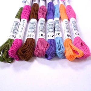 May include: Assortment of embroidery floss spools in various colors, including green, pink, brown, purple, blue, orange, and pink. The spools are labeled with numbers, including 392, 1110, 281, C 88, 112, 22, 02, and 07.