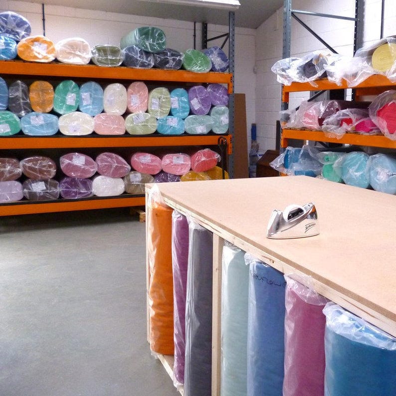 May include: A workshop with shelves full of colorful fabric rolls in various shades of pink, blue, green, and orange. The fabric rolls are wrapped in clear plastic and are stacked on orange shelves. A wooden table with a tape dispenser and a silver iron sits in the foreground. The table is also filled with fabric rolls in various colors, including orange, purple, gray, blue, and pink.