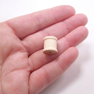 Puede incluir: Una pequeña bobina de madera sin terminar con un agujero en el centro. La bobina tiene un diámetro de aproximadamente 2,5 cm y una altura de 1,25 cm.