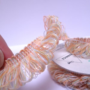 May include: A spool of peach, white and yellow fringe trim. The trim is made of a soft, delicate fabric and has a ruffled edge. The spool is labelled "Fabrics".