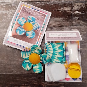 May include: A craft kit for making a flower brooch. The kit includes fabric, felt petals, felt circles, a brooch bar, and stuffing. The finished brooch is a blue and yellow flower with a geometric pattern. The packaging reads "FLOWER BROOCH" and "PAPER-AND-STRING."