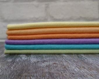 Puede incluir: Una pila de seis cuadrados de tela en colores pastel, incluyendo amarillo, naranja, morado claro y azul claro. Los cuadrados de tela están doblados cuidadosamente y dispuestos sobre una superficie de madera.