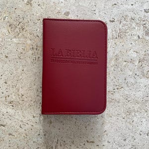 May include: A red Bible with the words "LA BIBLIA" and "TRADUCCIÓN DEL NUEVO MUNDO" embossed on the cover. The book has a smooth, leather-like texture and a rectangular shape. The background is a light-colored, textured surface.