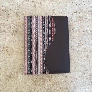 May include: A brown leather notebook cover with a pink and black patterned fabric insert. The fabric has a geometric design with stripes and zigzags.