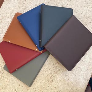 May include: A collection of seven leather-bound portfolios in various colors, including brown, red, blue, and gray. Each portfolio has a rectangular shape with rounded corners and a stitched border. The portfolios are arranged in a fan shape.