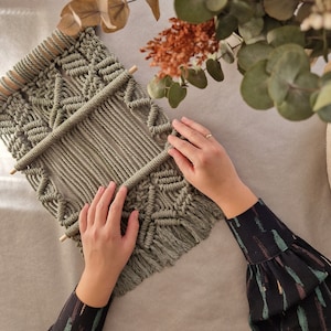 May include: A green macrame wall hanging with a geometric pattern. The wall hanging is made with natural fibres and has a fringe at the bottom.