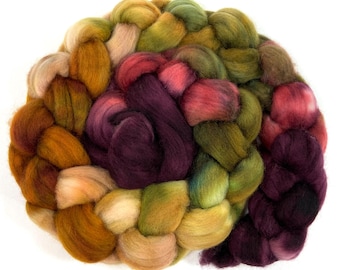 Puede incluir: Un paquete de vellón de lana trenzado en varios colores, incluyendo burdeos, verde, dorado y crema. La lana está dispuesta en forma circular, con los extremos de las trenzas superpuestos.