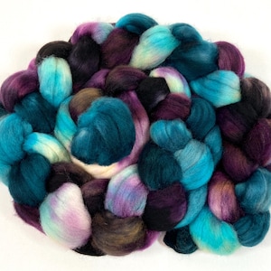 May include: A close-up of a ball of yarn with a mix of teal, purple, black, and white colors. The yarn is arranged in a loose braid.
