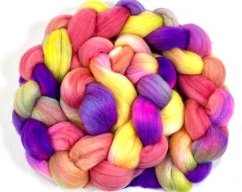 Può includere: Una colorata treccia di lana cardata tinta in tonalità rosa, gialle e viola. La fibra è disposta a cerchio, con ogni ciocca di lana che mostra una sfumatura di colori. La lana è morbida e soffice, ideale per progetti di fibre art.