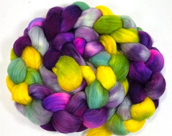 Può includere: Una colorata treccia di roving di lana nei toni del viola, giallo, verde e rosa. La fibra è fittamente intrecciata, creando una forma circolare strutturata. Il roving è ideale per filare, infeltrire o altri progetti di arte tessile.
